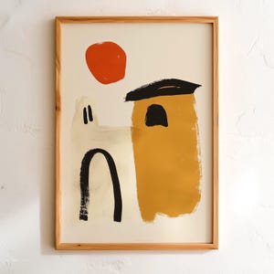 May include: A framed art print featuring an abstract illustration of two buildings in cream and mustard yellow with black arched doorways and a solid orange circle representing the sun, all enclosed within a light-toned wooden frame.
