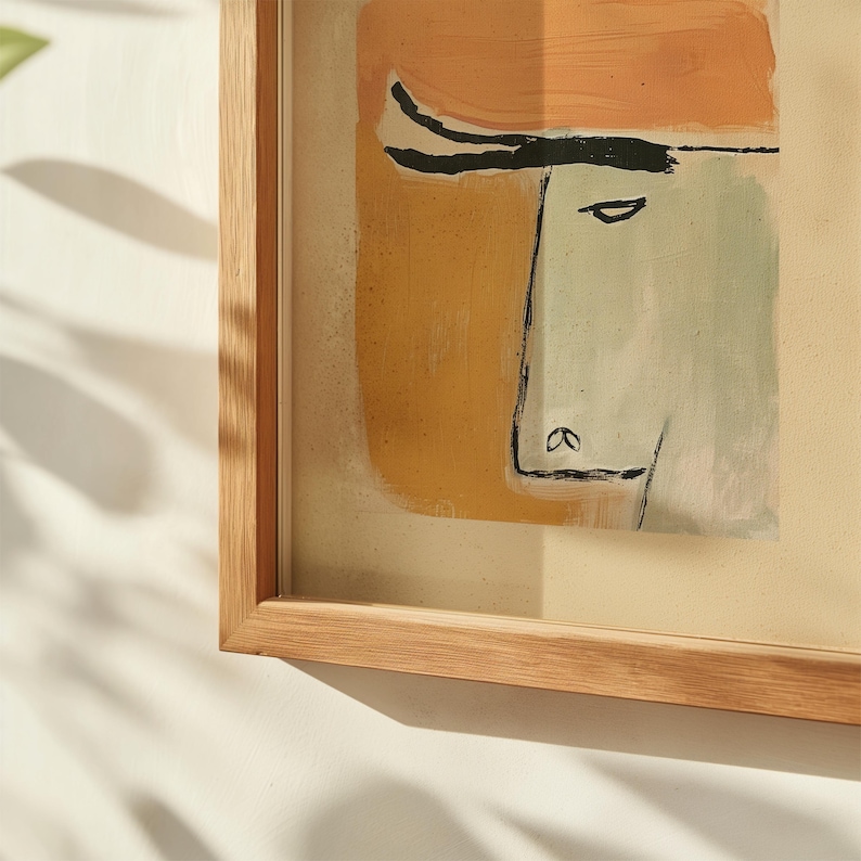 Op de afbeelding: Abstracte kunstprint met een minimalistisch gezicht met een lichtgroen gezicht en een oranje hoofdbedekking. Het kunstwerk is ingelijst in een natuurlijke houten lijst, wat een vleugje warmte toevoegt. Een uniek kunstwerk voor interieurdecoratie.