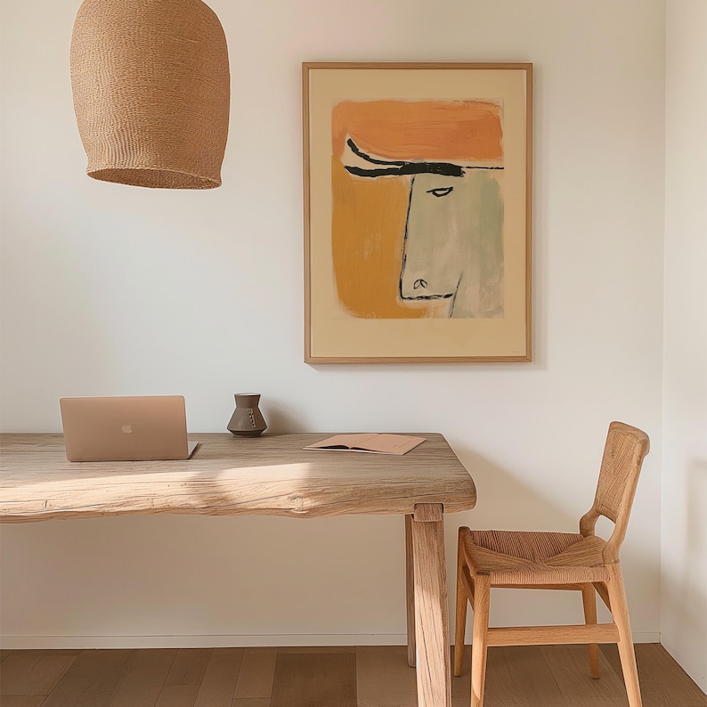 Op de afbeelding: Een lichte kamer met een houten bureau, een laptop, een kleine vaas en een ingelijst abstract kunstwerk. Boven het bureau hangt een geweven lamp. Rechts van het bureau staat een houten stoel met een geweven zitting.