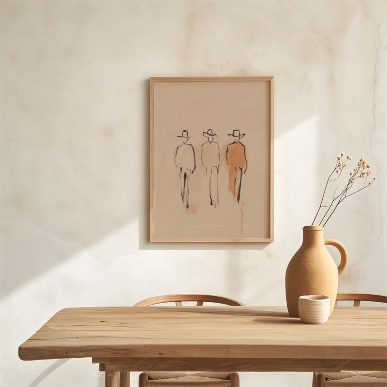 Puede incluir: Obra de arte enmarcada con un boceto minimalista de tres figuras con sombreros, en negro y naranja sobre fondo beige. La obra est&aacute; sobre una mesa de madera, con un jarr&oacute;n y una taza de cer&aacute;mica.