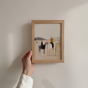May include: A framed art print featuring a minimalist illustration of two horses with riders. The artwork uses a neutral color palette with black, tan, and yellow accents. The frame is made of light-colored wood.