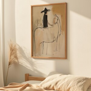 May include: Abstract art print in a wooden frame. The artwork depicts a person on a horse, rendered in black lines and beige tones. The person wears a black hat and shirt. The background has beige and tan brushstrokes.