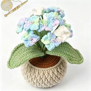 Op de afbeelding: Een gehaakte hortensia plant in een beige pot. De bloemen zijn pastelblauw, groen en paars, met parelaccenten. De bladeren zijn groen en de pot heeft een bruine binnenkant.