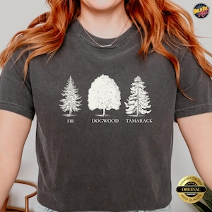 Può includere: T-shirt grigio scuro con tre illustrazioni di alberi bianchi: Abete, Corniolo e Larice. Il testo "FIR", "DOGWOOD" e "TAMARACK" è stampato sotto ogni albero. La maglietta ha un aspetto vintage.