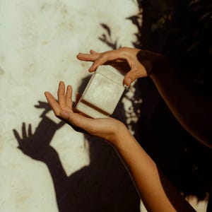 May include: A light-colored, rectangular box with a lid is held between two hands. The box appears to be made of stone or a similar material. The background is a textured, off-white wall with shadows.