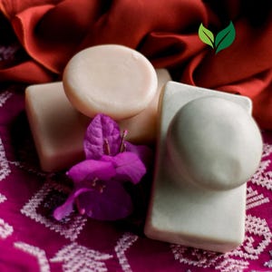 May include: Three bars of handmade soap, two are white and one is a light pink. The soap bars are on a red and white patterned fabric with a pink flower.