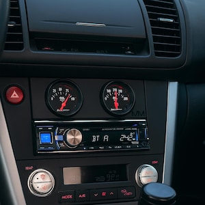 Puede incluir: Primer plano del salpicadero de un coche con indicadores, una radio y controles de climatización. Los indicadores muestran lecturas de aceite y temperatura. La radio tiene una pantalla digital. La palanca de cambios es visible. El salpicadero es negro y plateado.