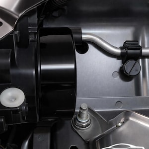 May include: Close-up of a car engine compartment with various components. The image features a black cylindrical part, a silver pipe, and several bolts. The background is a metallic gray color, with the CarCultureCyprus logo visible.