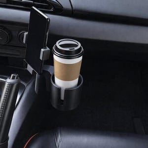 Puede incluir: Interior de coche negro con un soporte para teléfono y un portavasos. Una taza de café desechable con tapa negra está en el portavasos. Un teléfono inteligente está en el soporte del teléfono. El asiento del coche es negro.