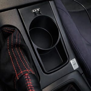 May include: A black automotive center console cup holder featuring a silver GT-R emblem, a circular cup slot, and a rectangular storage compartment. Adjacent is a black gear shift boot with red stitching and a small gear shift pattern decal.