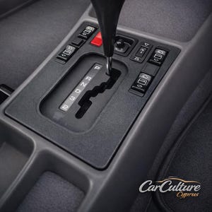 May include: Close-up of a car's interior featuring a black gear shift lever and console. The gear selector is labeled P, R, N, D, 3, and 2. The console also has buttons and a red hazard light button. The car's interior is gray.