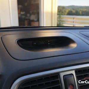 May include: A close-up of a car dashboard with a digital clock displaying 7:02. The dashboard is black with a textured surface and a recessed area for the clock. The image is taken in daylight.