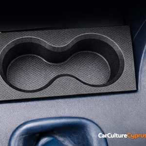 May include: Black car interior console featuring a cup holder and a cigarette lighter port. The cup holder has two circular openings. The console is set in a blue interior.