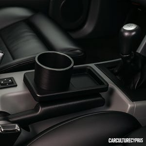 May include: Black cup holder and tray accessory for a car's center console. The cup holder is cylindrical, and the tray is rectangular. The interior of the car has black leather seats and a gray console. A seat belt buckle is visible.