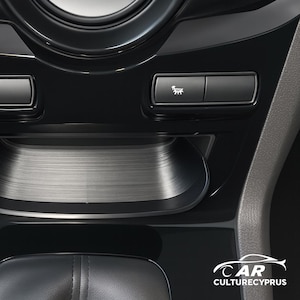 Può includere: Primo piano del cruscotto di un'auto con finitura nera lucida. Include un vassoio in metallo spazzolato, due pulsanti rettangolari e un pulsante con un'icona di auto. L'interno è elegante e moderno.