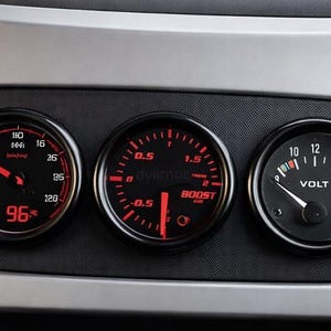 Puede incluir: Indicadores de salpicadero con esferas negras y agujas rojas. Los indicadores incluyen un velocímetro que marca 96, un indicador de sobrealimentación y un voltímetro. Los indicadores están colocados en un salpicadero plateado y negro.