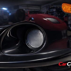 May include: Close-up of a black car's front bumper, featuring a fog light with a silver mesh cover and an amber turn signal. The car's glossy finish reflects the surrounding environment.