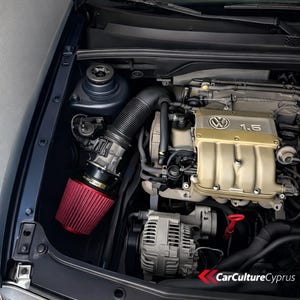 Puede incluir: Compartimento del motor de un coche con un filtro de aire rojo y un tubo de admisión negro. El motor tiene una cubierta dorada con el logotipo de Volkswagen y el texto "1.6". El compartimento del motor es azul oscuro.