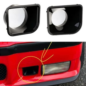 May include: Two black rectangular automotive air duct inserts with rounded corners and a smooth finish. Below, a red car bumper features a matching black air duct opening highlighted by a yellow circle and arrow, positioned next to a clear fog light.