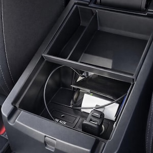 Può includere: Console per auto nera con più scomparti, tra cui una porta di ricarica e un ingresso AUX. Un cavo di ricarica e uno smartphone sono visibili all'interno. Interno di un'auto.