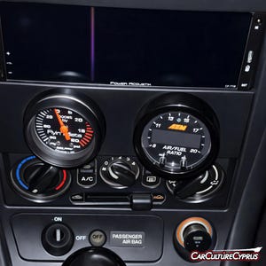 Op de afbeelding: Zwart auto dashboard met twee ronde meters en een digitaal display. De linker meter geeft de turbodruk aan. De rechter meter toont de lucht/brandstofverhouding. Het display toont "Power Acoustik".