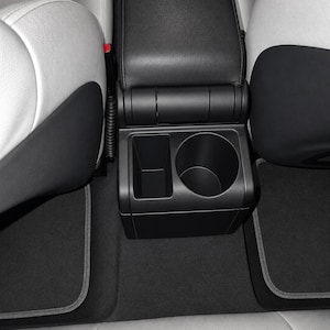 May include: A black plastic center console with a rectangular storage compartment and a circular cup holder, positioned between two light gray and black car seats with dark gray floor mats featuring light gray piping on the floor below.