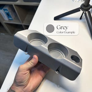 May include: A grey, rectangular, 3D-printed plastic cup holder insert featuring two large circular openings and one smaller circular opening on the side. The surface has a smooth, matte finish with visible mounting tabs along the bottom edge.