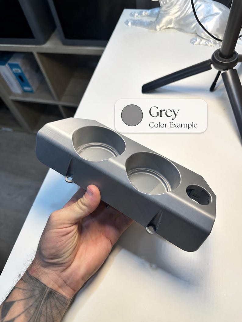May include: A grey, rectangular, 3D-printed plastic cup holder insert featuring two large circular openings and one smaller circular opening on the side. The surface has a smooth, matte finish with visible mounting tabs along the bottom edge.