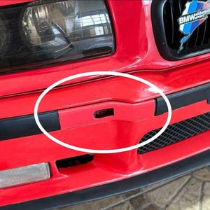 May include: A close-up of a red car bumper with a black accent. The bumper features a rectangular opening and is part of a vehicle's front end. The image highlights the car's design and colour.