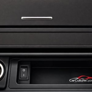以下が含まれることがあります： 黒い車のダッシュボードのクローズアップ。シルバーのアクセント、DSCボタン、電源コンセント、スイッチが含まれています。CarCultureCyprusのロゴが赤と白で表示されています。