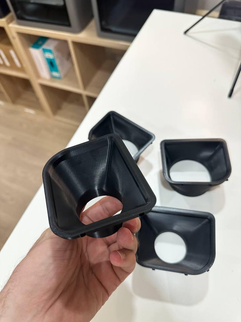 May include: A set of black, 3D-printed plastic funnel-shaped components with a square-to-round transition design. Each piece features a wide, square-shaped opening that tapers down into a circular aperture, showcasing a smooth, matte finish.