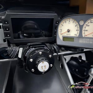 May include: Close-up of a black automotive component with a circular design and silver accents. The component is mounted near a dashboard with gauges and a screen. The text "125HH040105" is visible on the component.