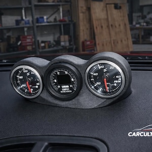Op de afbeelding: Een zwart dashboardmetercluster met drie ronde meters. De meters hebben witte cijfers en rode naalden. De middelste meter heeft een digitaal display. Het dashboard is zwart.