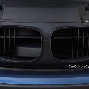 May include: A close-up shot of a black car grille featuring vertical bars and a dual kidney shape. The grille is set against a dark blue car body. The text "CarCultureCy" is visible in the lower right corner.