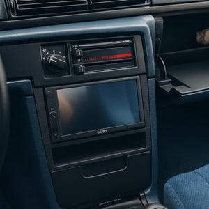 Può includere: Vista interna del cruscotto di un'auto d'epoca. Il cruscotto è blu scuro e presenta un display touchscreen Sony nero, bocchette dell'aria e una radio. Sono visibili anche un cambio nero e un sedile in tessuto blu.