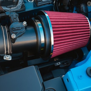 May include: A detailed view of a car engine bay featuring a red air filter. The filter, with a chrome trim, is connected to black tubing. The engine components are set against a blue background. The image includes the text "CAR CULTURE CYPRUS".