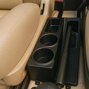 May include: A black plastic center console organizer featuring two circular cup holders and a rectangular storage compartment, positioned between beige car seats next to a manual gear shift lever and a handbrake with a red release button.