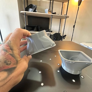 May include: Two light gray, 3D-printed plastic components with a flared, funnel-like shape. Each piece features a square opening at the top that tapers down to a circular base, with small, evenly spaced tabs protruding along the square rim edge.