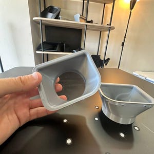 May include: Two grey, 3D-printed plastic funnel-shaped components with a square-to-round transition design. Each piece features a wide, square-shaped opening that tapers down into a circular aperture, showcasing visible layer lines from the manufacturing process.