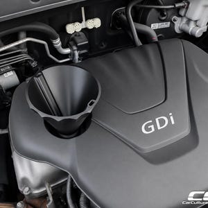 May include: A black plastic funnel inserted into the oil filler neck of a car engine. The engine cover is matte black with the silver text GDi printed on the top. Various metal components, hoses, and electrical connectors are visible in the background.