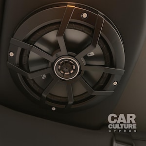 Può includere: Un altoparlante audio per auto nero con una griglia protettiva, montato su una superficie scura. L'altoparlante ha un emblema centrale ed è circondato da una cornice circolare. L'immagine mostra anche una parte di un apparecchio di illuminazione.