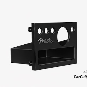 May include: Black car dashboard component with the word "Miata" in a cursive font. The component has three small circular holes, one large circular hole, and a rectangular opening. The component is designed for a car.