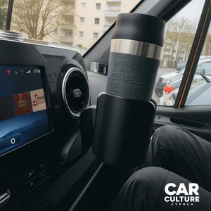 Può includere: Un bicchiere da viaggio Stanley nero e argento in un portabicchieri per auto. Il bicchiere ha la scritta "STANLEY" e "NONVESIDE" stampata sopra. Sono visibili il cruscotto dell'auto e una parte delle gambe del conducente.