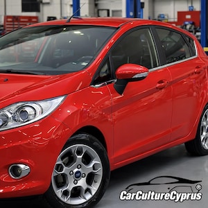 May include: Bright red Ford Fiesta sedan with silver alloy wheels. The car has a sleek, modern design with a glossy finish. The image is taken in a well-lit garage setting.
