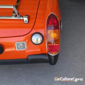 Puede incluir: Primer plano de la parte trasera de un coche clásico naranja. La imagen destaca el piloto trasero, una luz redonda plateada y una luz rectangular. Se ve un portaequipajes cromado. La carrocería es de color naranja brillante.