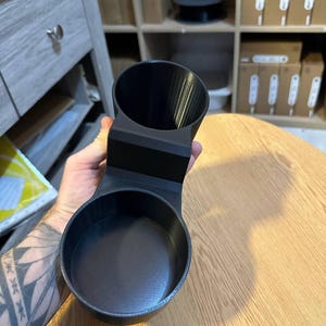 May include: A matte black 3D printed dual cup holder accessory featuring two circular openings of different sizes connected by a central bridge, designed with a smooth interior finish and a textured exterior surface for secure placement.