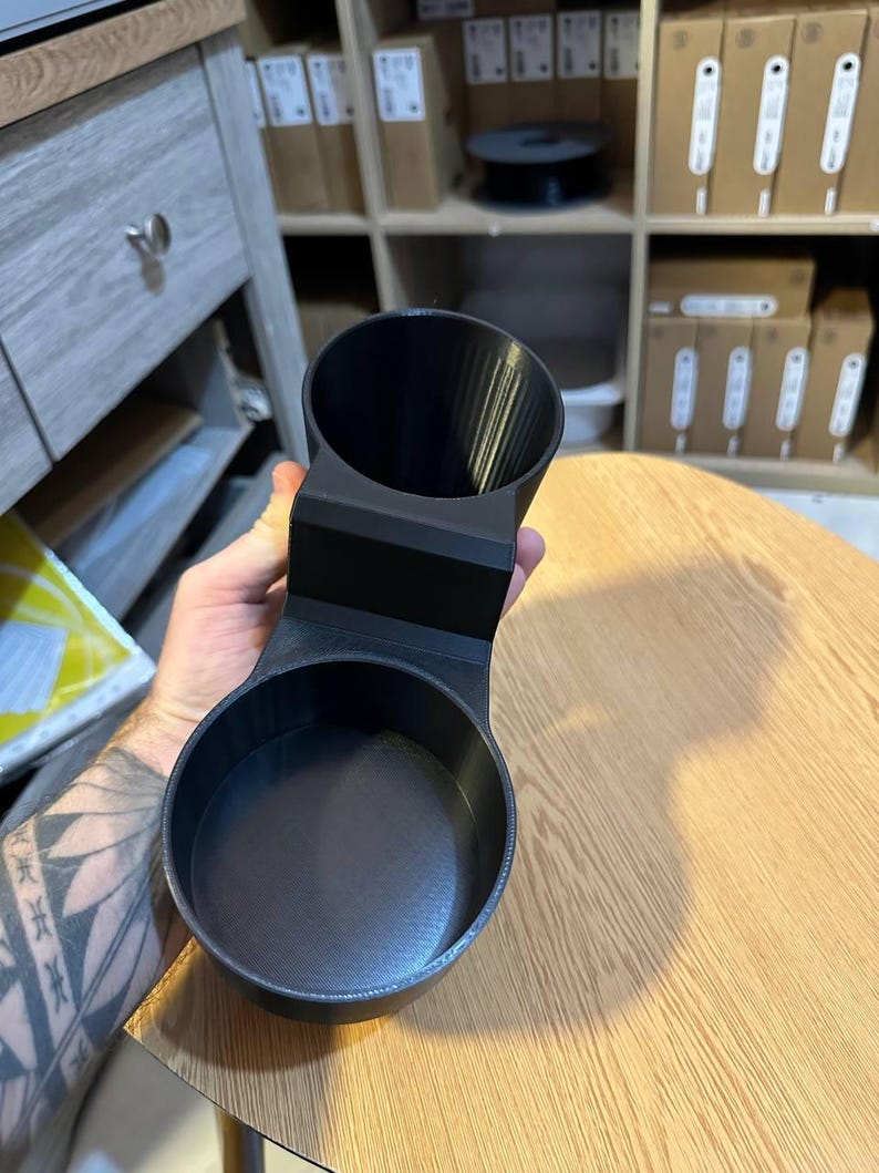 May include: A matte black 3D printed dual cup holder accessory featuring two circular openings of different sizes connected by a central bridge, designed with a smooth interior finish and a textured exterior surface for secure placement.