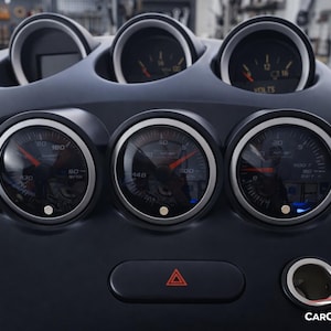 Könnte beinhalten: Schwarzes Armaturenbrett mit mehreren Messgeräten und Zifferblättern. Die Messgeräte haben silberne Ränder und rote Nadeln. Das Armaturenbrett verfügt außerdem über eine Warnblinkertaste und einen Zigarettenanzünderanschluss. Der Text "VOLTS" ist auf einem der Messgeräte sichtbar.