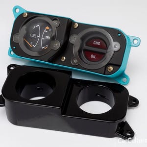 Può includere: Un gruppo di indicatori automobilistici verde acqua e neri con due indicatori, uno che mostra i livelli di carburante e temperatura e l'altro che mostra gli indicatori di carica e olio. Sotto, un alloggiamento nero lucido a doppio foro.
