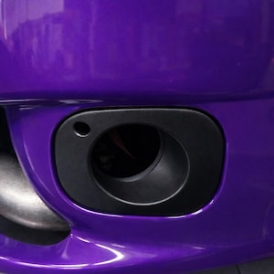 Może przedstawiać: Zbliżenie fragmentu przedniego zderzaka samochodu. Zderzak jest w żywym fioletowym kolorze z błyszczącym wykończeniem. Widoczna jest czarna, owalna obudowa światła przeciwmgielnego. Logo Car Culture Cyprus znajduje się w prawym dolnym rogu.